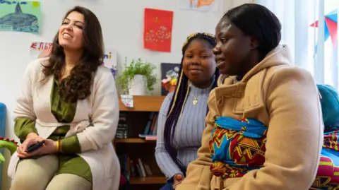 International Rescue Committee 3 women sitting in a room, smiling. The walls behind them are decorated with colourful children's drawings. 