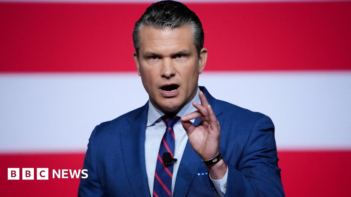 Hegseth in blue jacket and red and blue striped tie with his mouth open as he speaks stands in front of a background of large horizontal red and white stripes with his left hand raised, thumb and forefinger pressing together