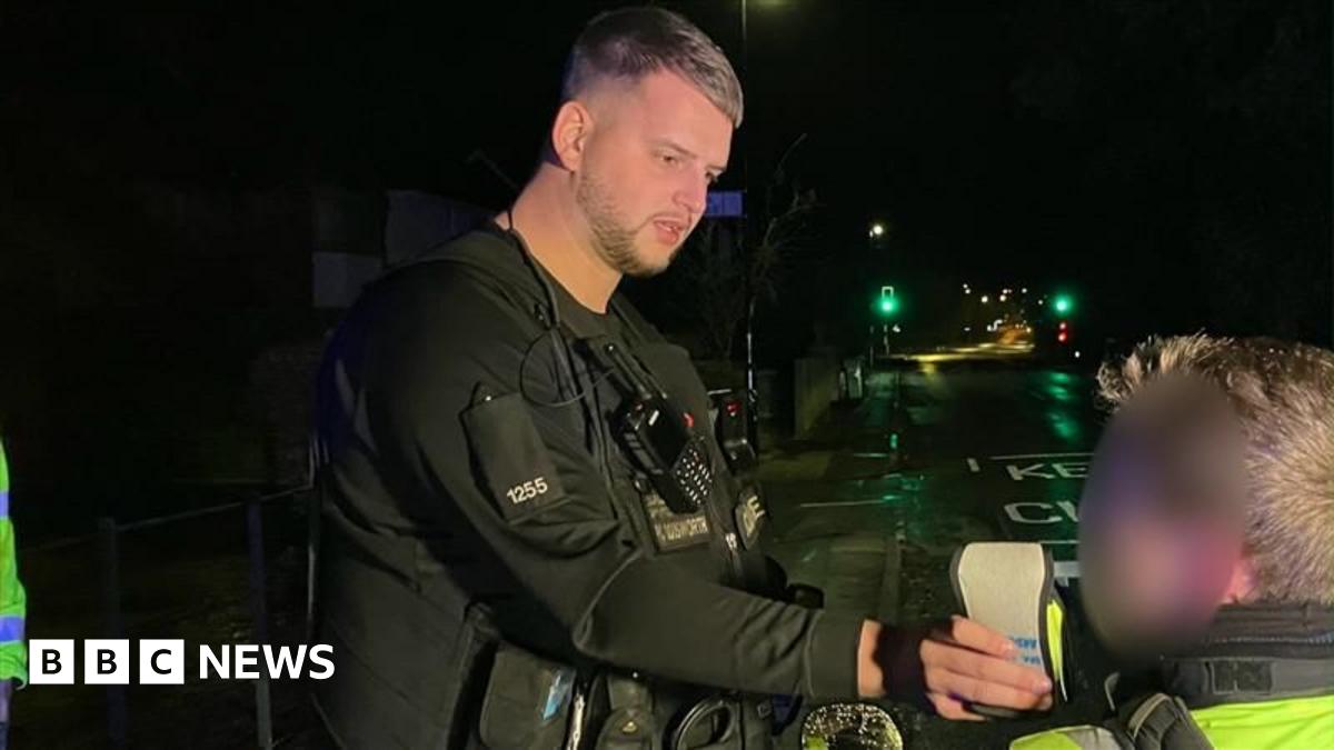 A traffic officer dressed in black uniform holds out a breathalyser device while a young man, whose face is blurred, blows into it on a dark street.