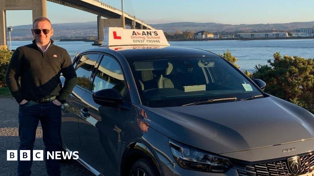 A dark grey car with a learner driver sign reading 'Alan's Driving School' parked near a waterfront, with a large suspension bridge in the background and hills visible across the water