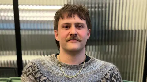 Jordan Garland has short, brown hair and a moustache and is sitting in front of a distorted glass window. He is wearing a gold chain and a grey jumper with blue and black geometric prints