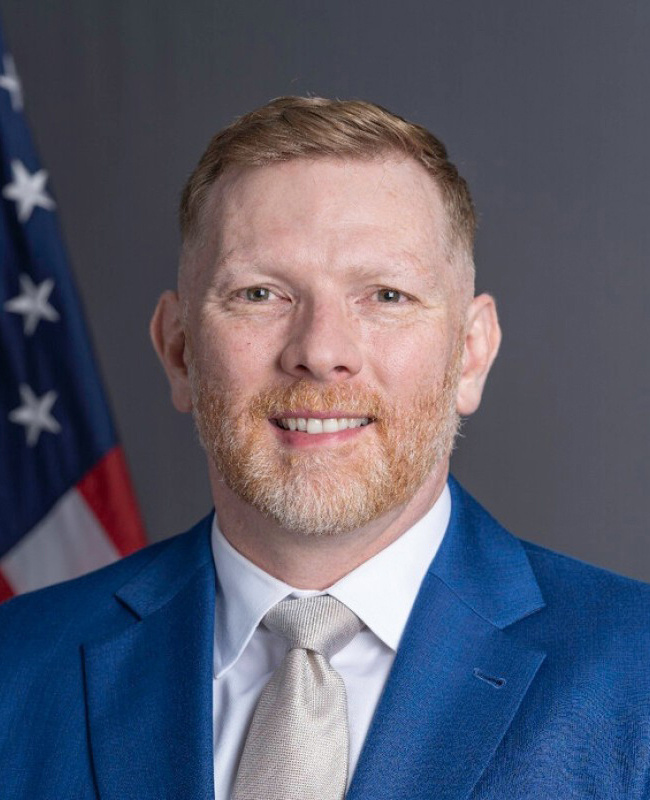 A headshot of a man in a suit looking at the camera, in front of the American flag.