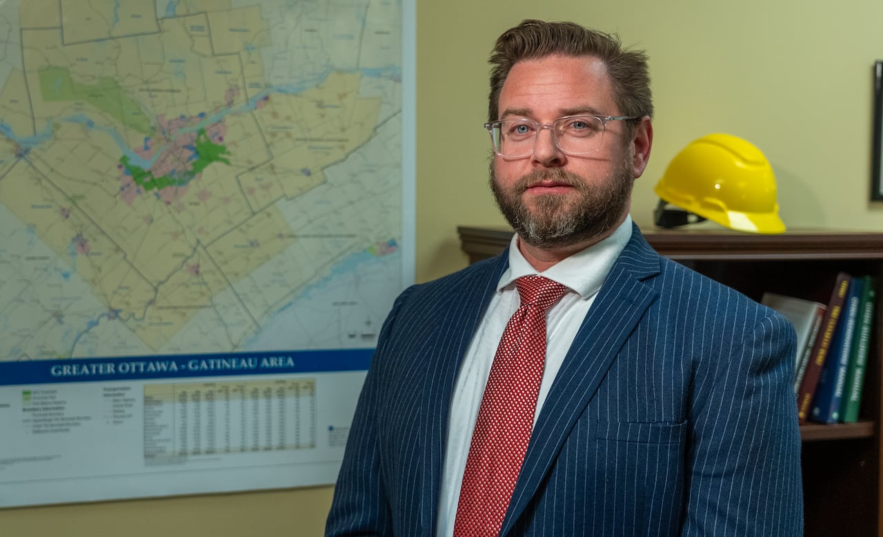 A man with glasses stands in front of a map of Ottawa and construction hat.