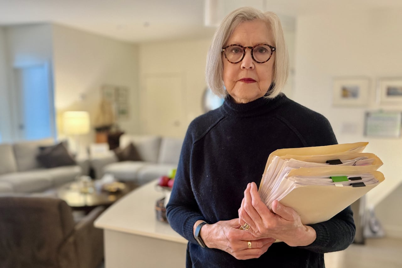 A woman wearing a dark sweater stands is standing in her apartment holding three folders filled with documents.
