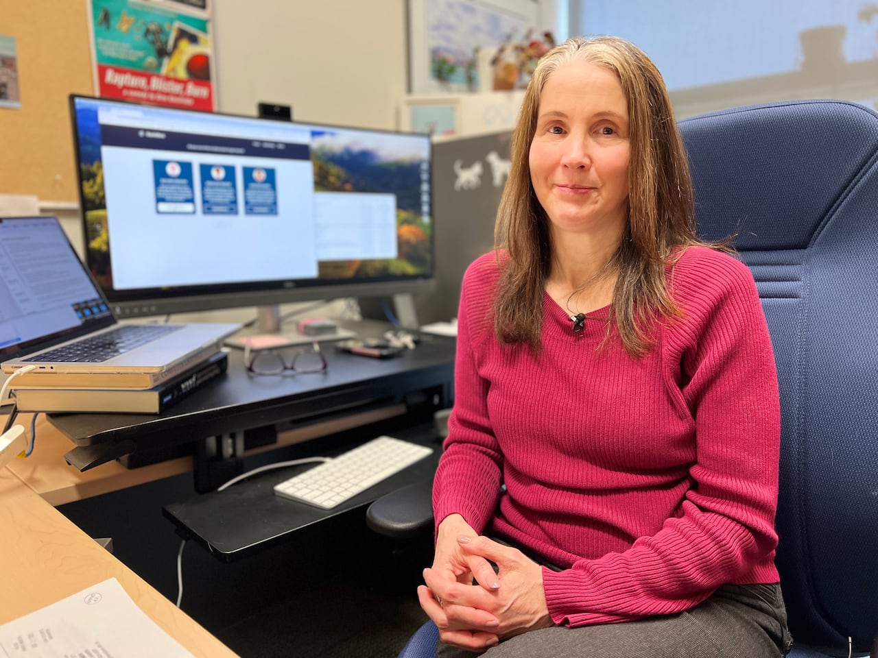 A woman is seated in a chair beside a desk with a laptop computer and a desktop computer screen and keyboard.