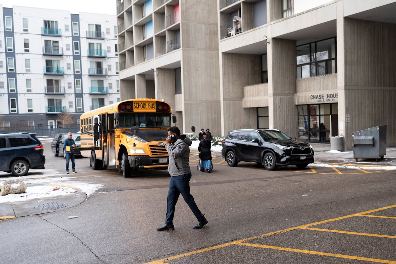 A man on a cell phone walks past an apartment complex with a school bus parked out front