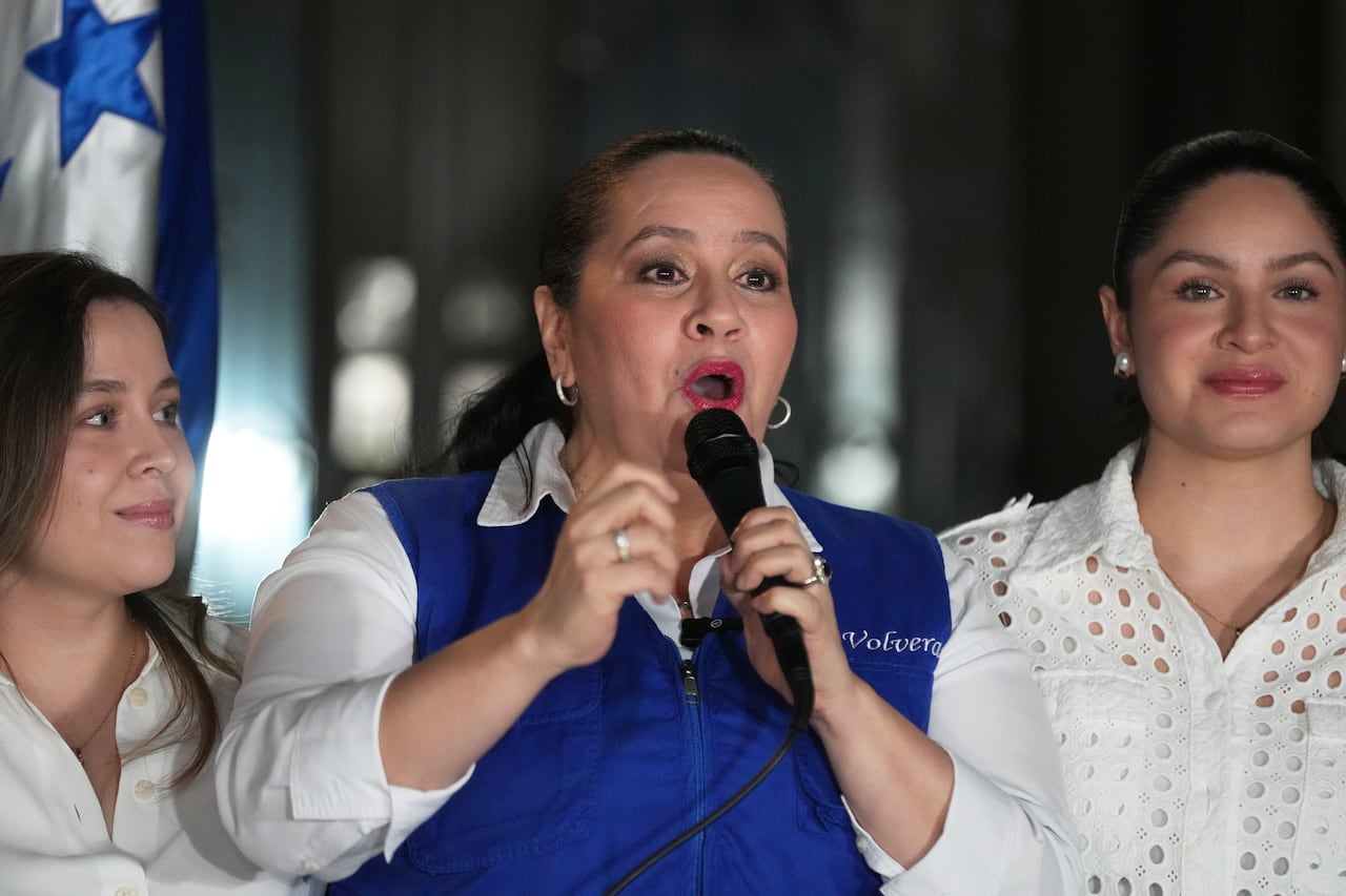 A dark haired woman with her hair tied back, speaks into a microphone while surrounded by two younger women on either side.