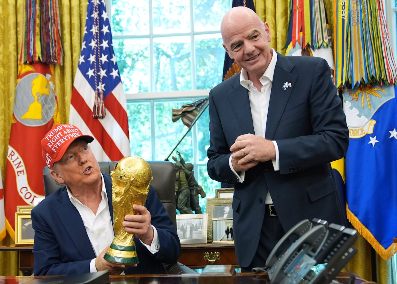 Donald Trump sits at a desk wearing a red hat that says 'Trump was right about everything' as he holds a golden trophy. A smiling bald man in a suit stands next to him.