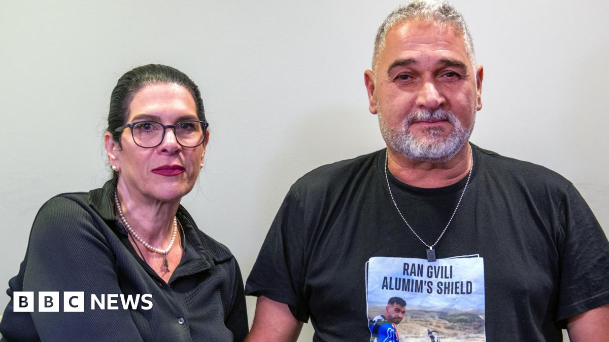Talik, who has black hair in a ponytail and wears black-rimmed glasses, poses for a photo with her husband Itzik, who has grey short hair and a beard and wears a silver chain. They both have neutral expressions.