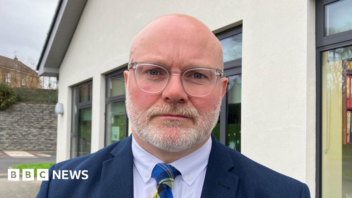 A bald man with glasses and a white beard is standing in front of a school building during the day. He is wearing a blue suit, shirt and blue and yellow pattern tie.