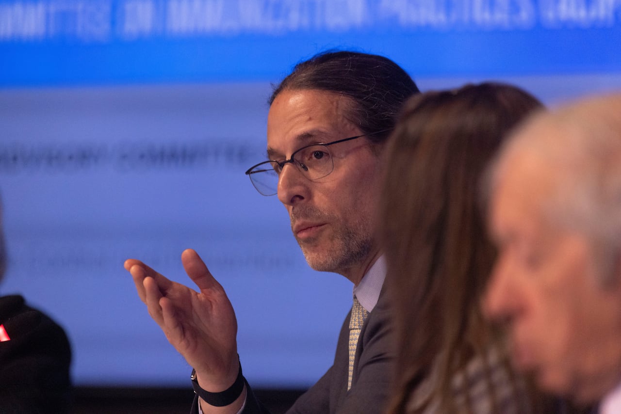 a man with glasses and a ponytail in a suit speaks to a group of others on a panel