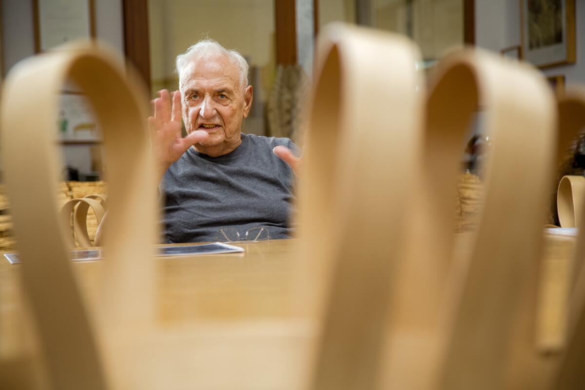 Frank Gehry is seen through looping wood forms as he talks, gesturing with his hands.