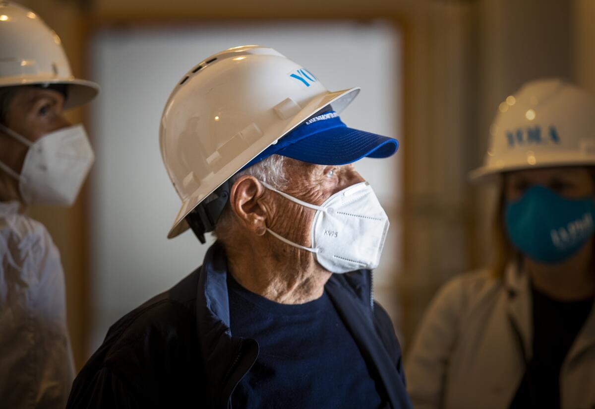 Frank Gehry wears a face mask and a construction helmet as he tours a construction site.