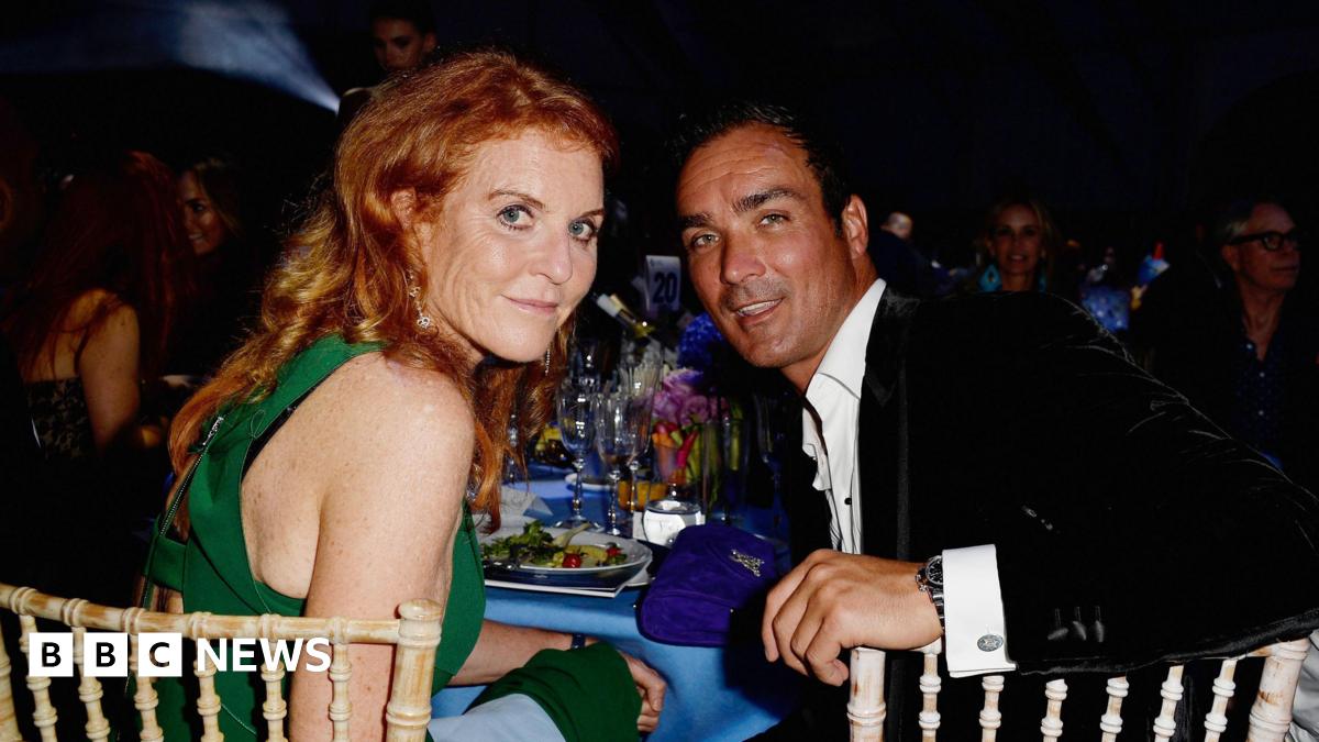 Sarah Ferguson and Manuel Fernandez sit at a table laden with glasses, plates and a blue table cloth. She wears a green dress, he wears a black velvet jacket and a white shirt open at the neck. They sit at wickerwork-style chairs, and are looking over their shoulders at the camera