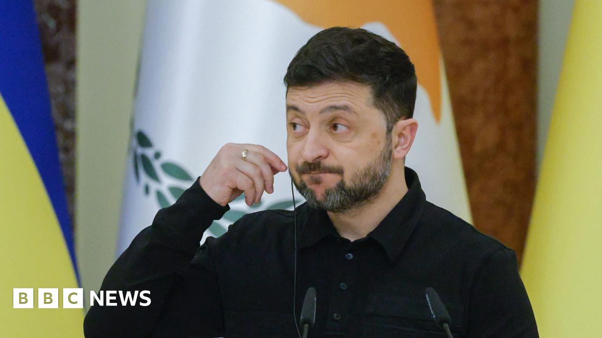 The Ukrainian president, bearded and dressed in black, checking his mic and stood in front of some  flags