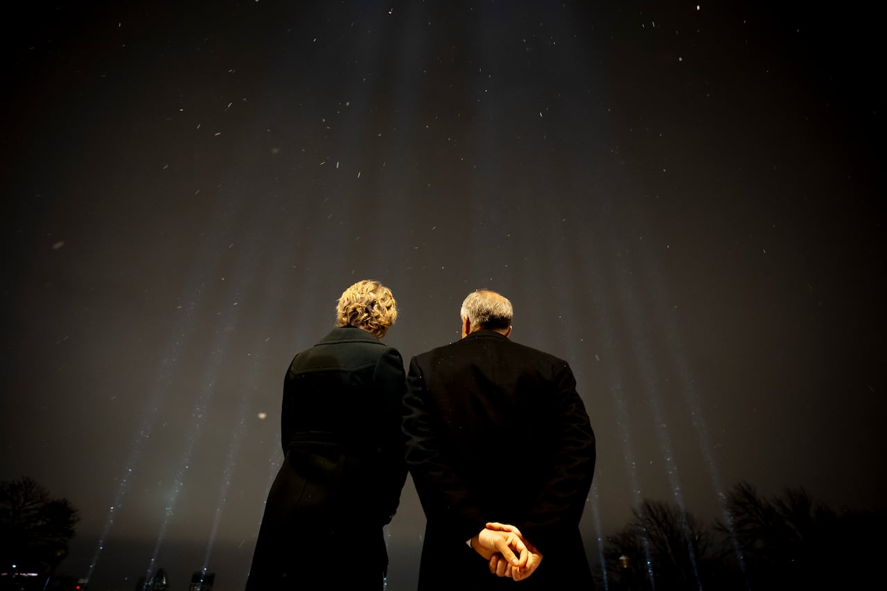 A man and a woman are seen with their backs to the camera looking up at 14 beams of light pointing skyward in the night.