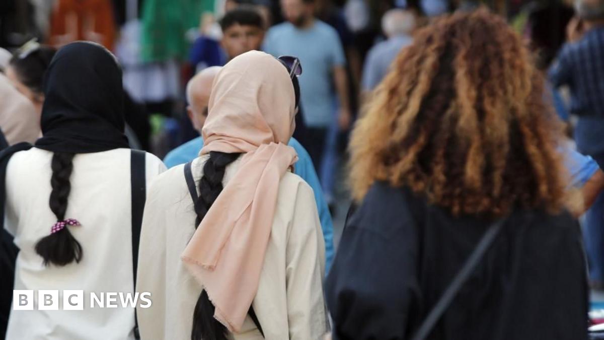 Three Iranian women, one wearing a black head covering, another with a pink hijab and  a fourth one without a head covering with thick curly brown hair, walking.