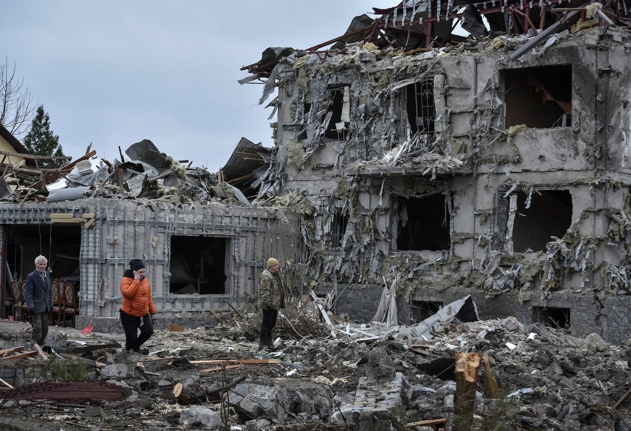People stand near a ruined building and amid rubble 
