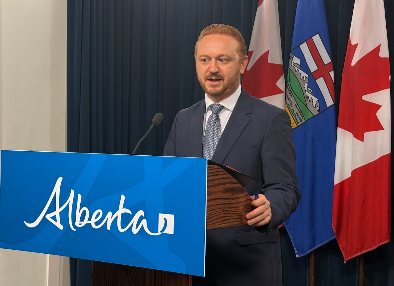 A man with red hair stands behind a podium with the words "Alberta" on the front, with the Canada and Alberta flags behind him.