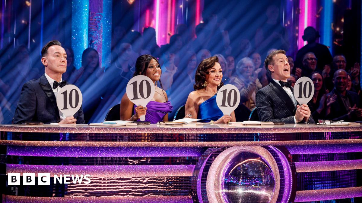 George Clarke wears a blue and white top with purple trousers and dances next to his partner Alexis Warr who wears an orange two piece with frills, they have their hands on their head as they dance and there are three backing dancers behind them.