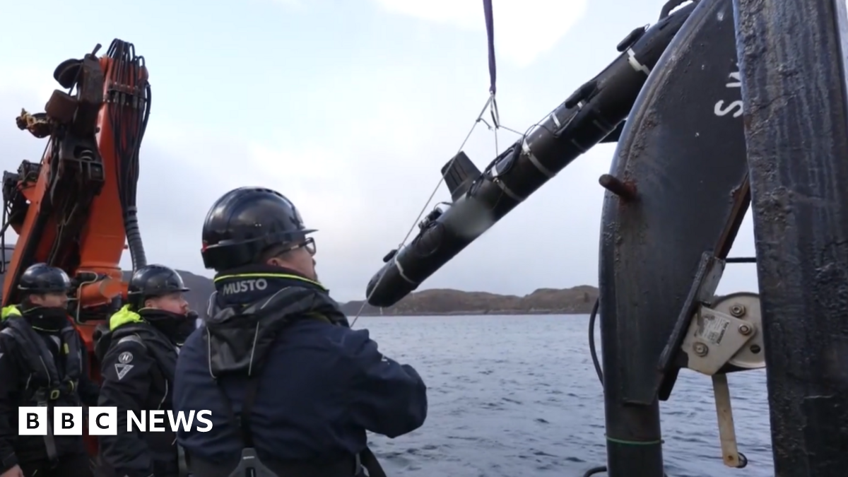 Three men in hardhats onboard a ship launching  a SG-1 Fathom glider into the sea. It is lifted by a pulley and lowered into the water
