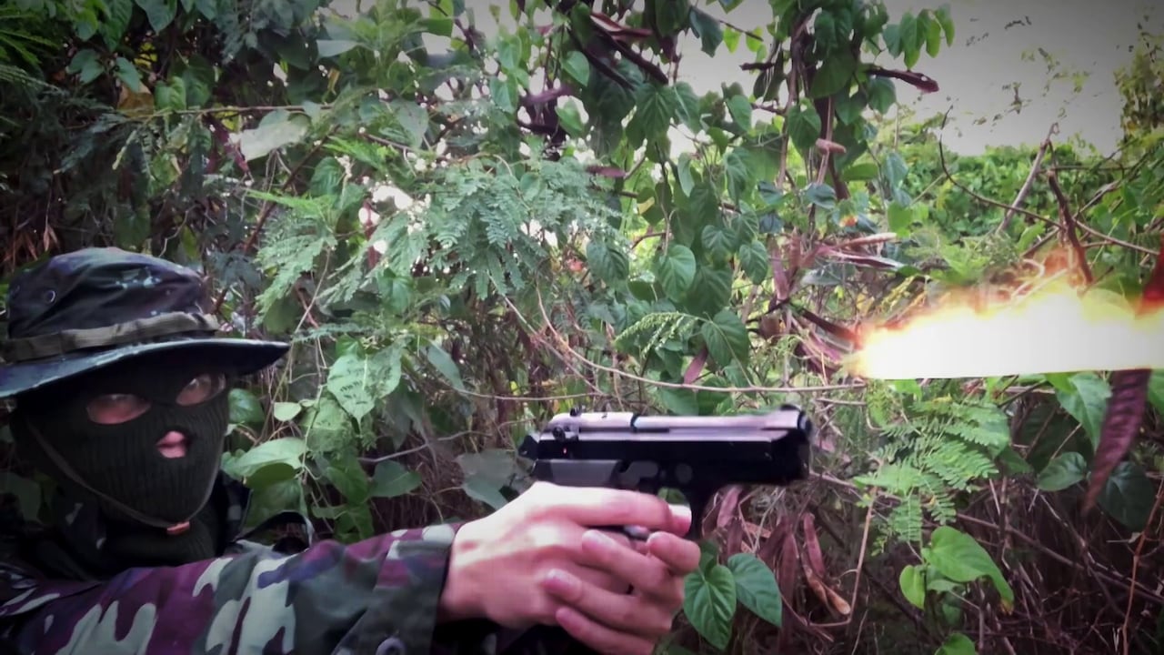 A man in a three-hole balaclava fires a blank in a jungle setting.