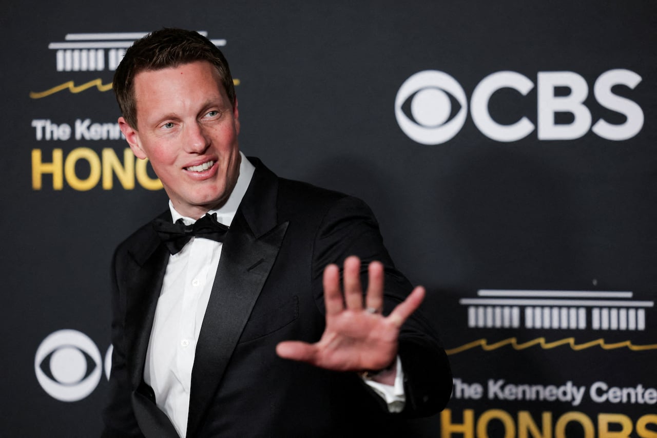 A cleanshaven man in a dark tuxedo and bow tie is shown waving to someone off camera. 