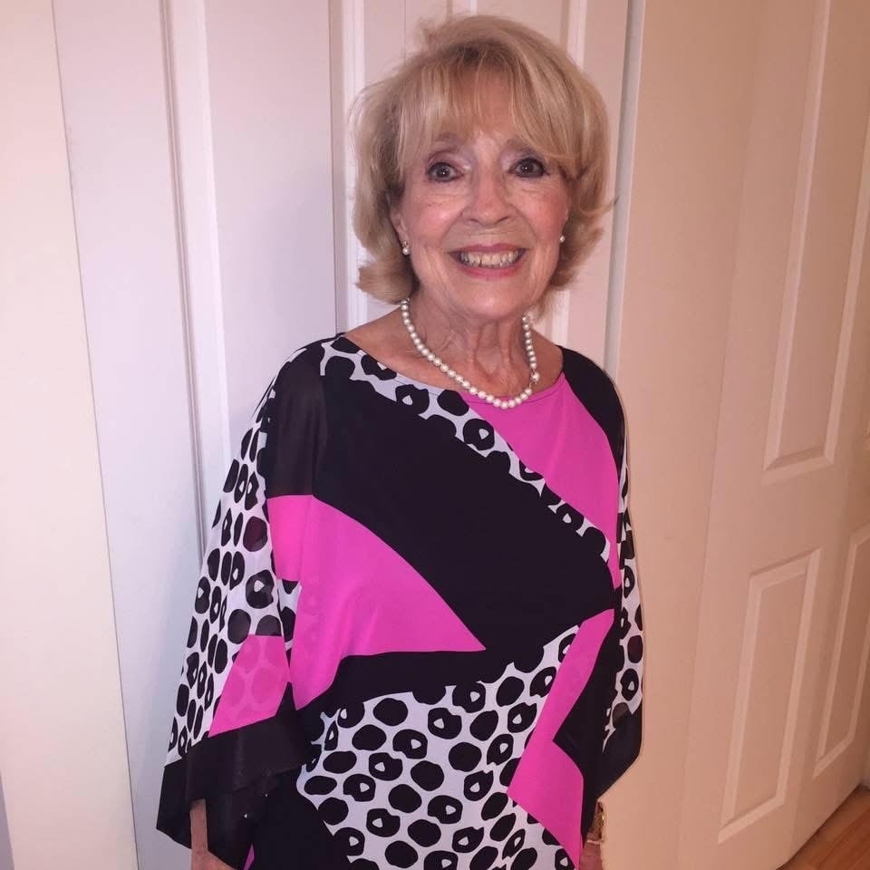 Blond elderly woman wearing black, white and pink dress standing in front of a white door. 
