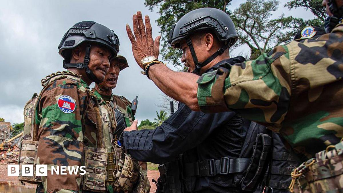 Two troops in Cambodian military uniform and helmets face off with a troop in black uniform and helmet, while in the foreground another soldier in military uniform holds out his hand with the palm acing outwards