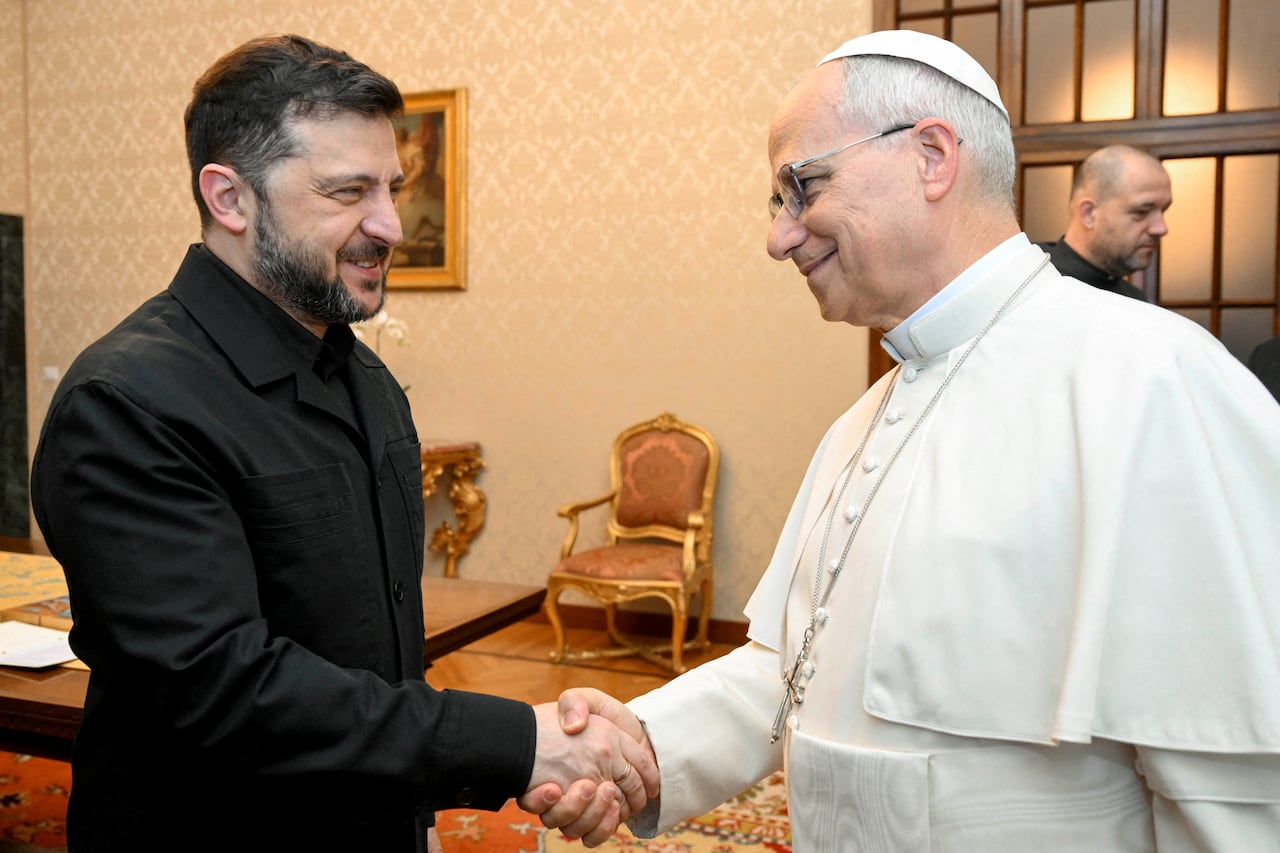 A bearded man in dark clothing shakes hands with the pope, who is bespectacled, wearing a skull cap and dressed in white.