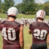 Two players with their backs to the camera watch the players on the football field from the sidelines. One is wearing jersey No. 40 and one wears No. 20. Both are wearing their football helmets. 
