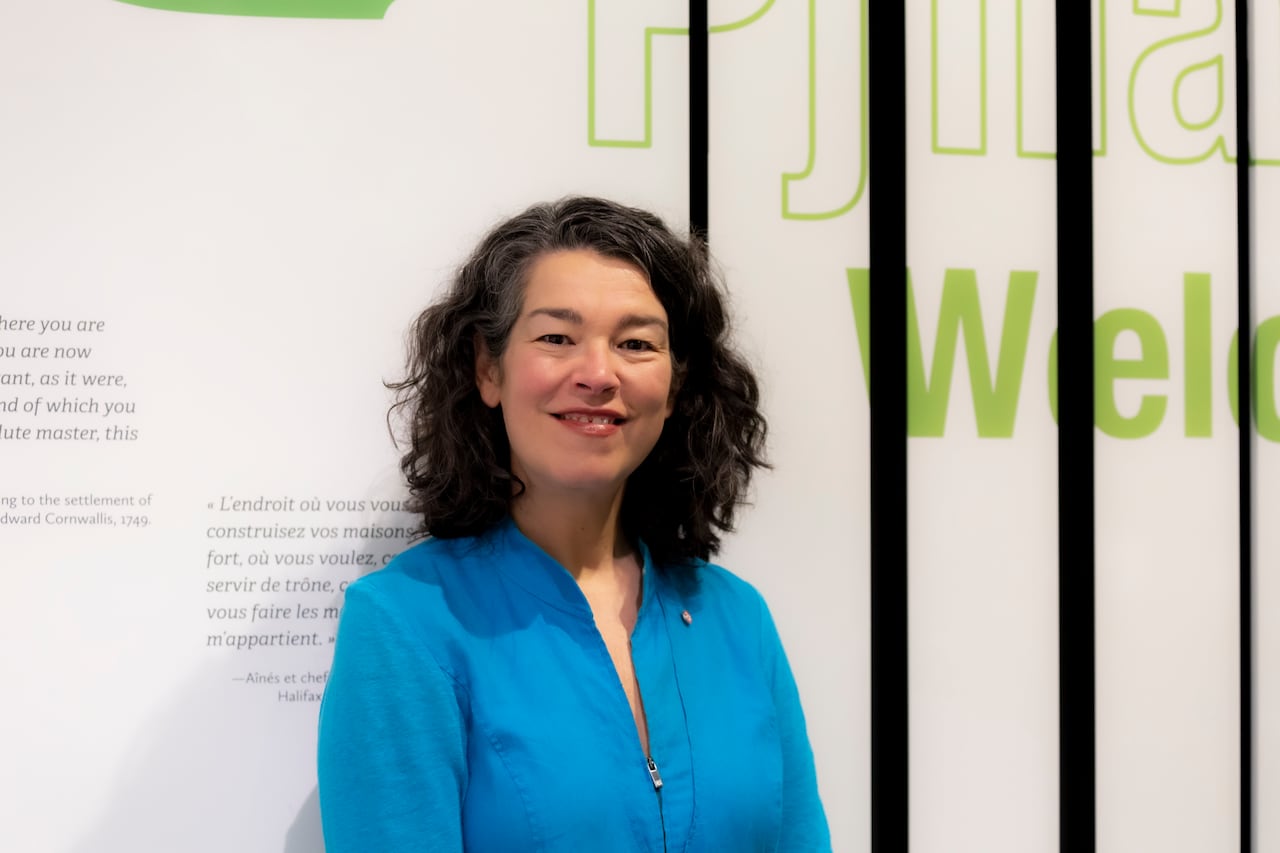 A woman with shoulder-length wavy dark hair wearing an aqua-coloured blouse with a small zipper stands in front of a white exhibit wall with lettering on it.