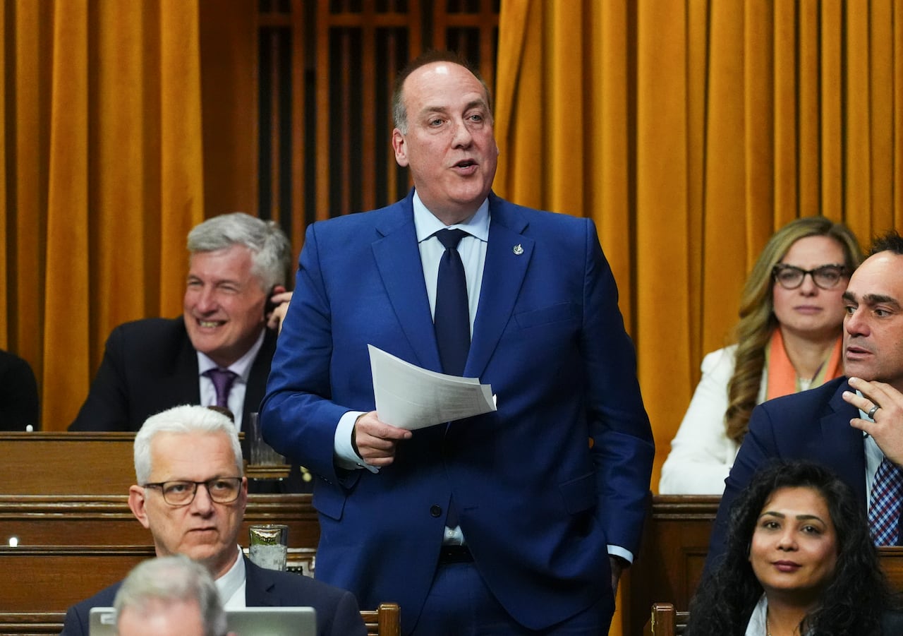 A man in a blue suit stands and speaks while holding a piece of paper.