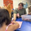 A little girl with her back to the camera sits and draws at a low table the color of blueberries in a classroom. Her preschool teacher leans in with a crayon to help. A plastic bin containing crayons sits between them.