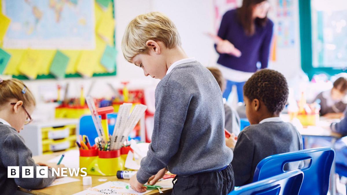 A blonde haired boy in a grey uniform stands at a desk doing a craft. Some other students are sitting at a table. A teacher at the top of the class wearing a purple top looks at some of their work.