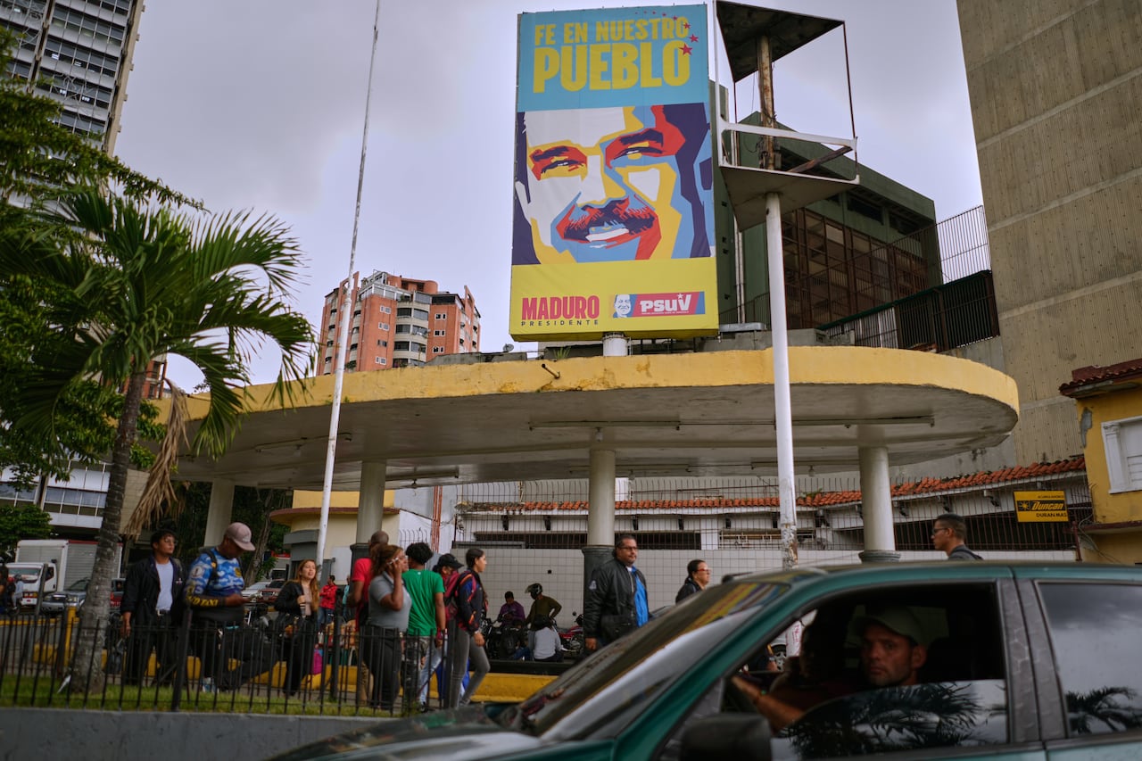 A crowd of people standing under a roofed structure, with a poster of Nicolás Maduro mounted above it.