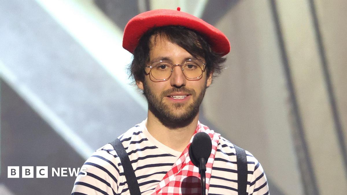 A man in a red beret and striped t-shirt stands on stage behind two microphones as he delivers a speech.