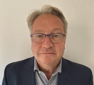 Man wearing glasses, dark jacket and light grey shirt stands in front of white background. 