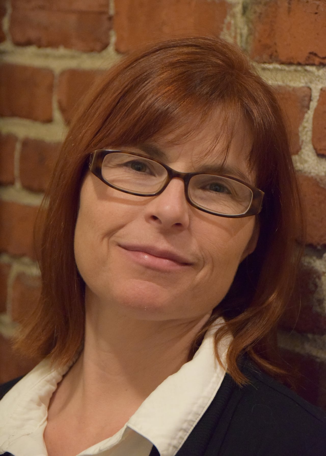 Woman wearing glasses, white shirt and a black sweater who is standing in front of brick wall