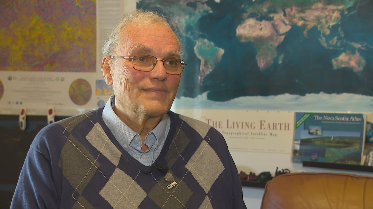 A man a collared shirt and sweater, sitting in front of a poster with a map of the earth.