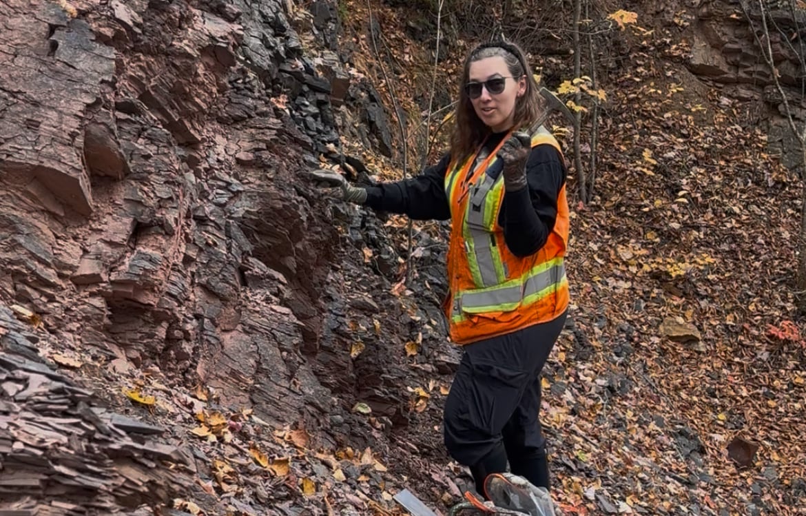 A woman in a safety vest stands near a rock face holding a hammer.