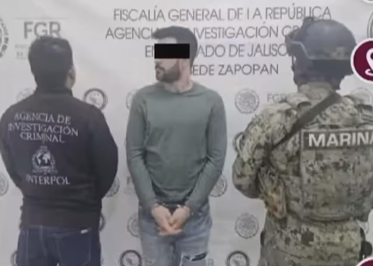 A man in handcuffs, between a man in an INTERPOL jacket and another man in camouflage.