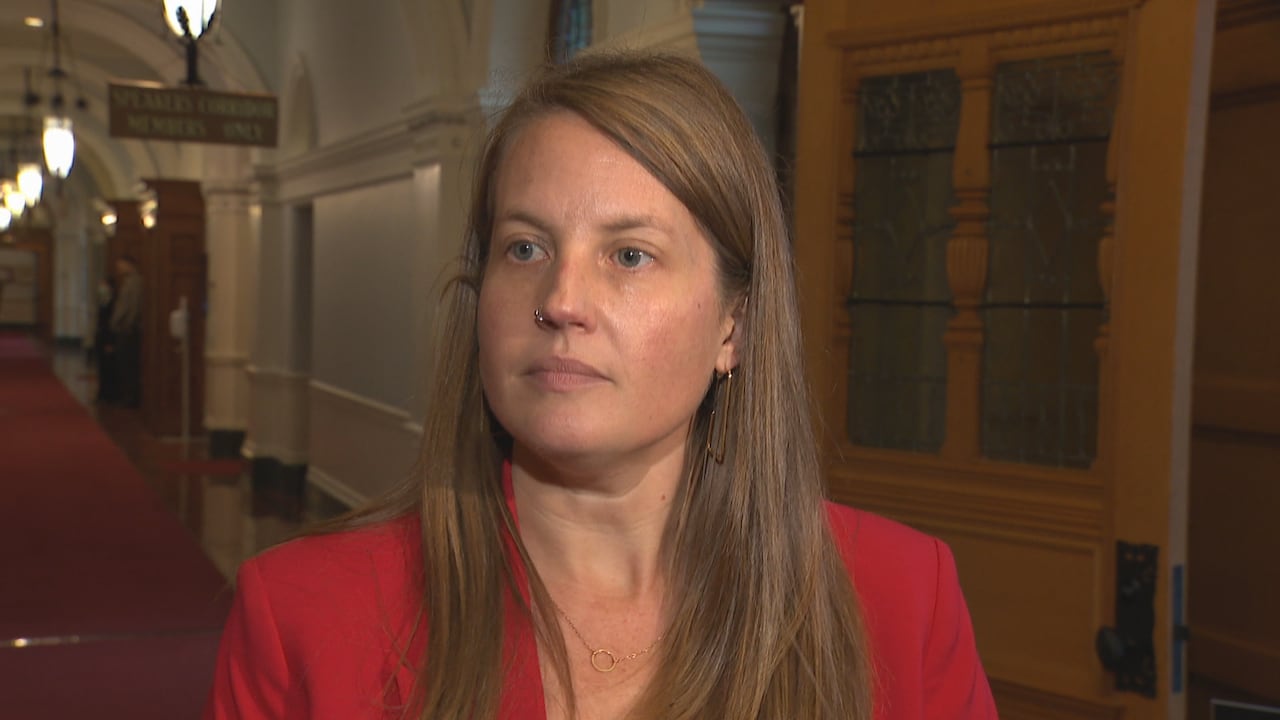 A woman in a red blazer speaks in a legislature hallway