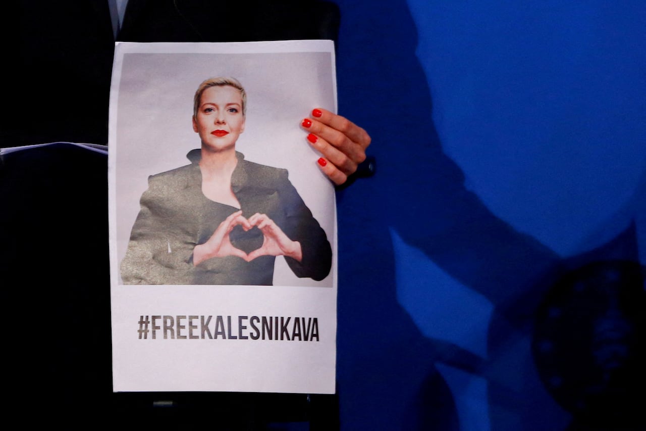 A woman holds up a poster that has a picture of another woman with the words #FreeKalenikava written underneath. 