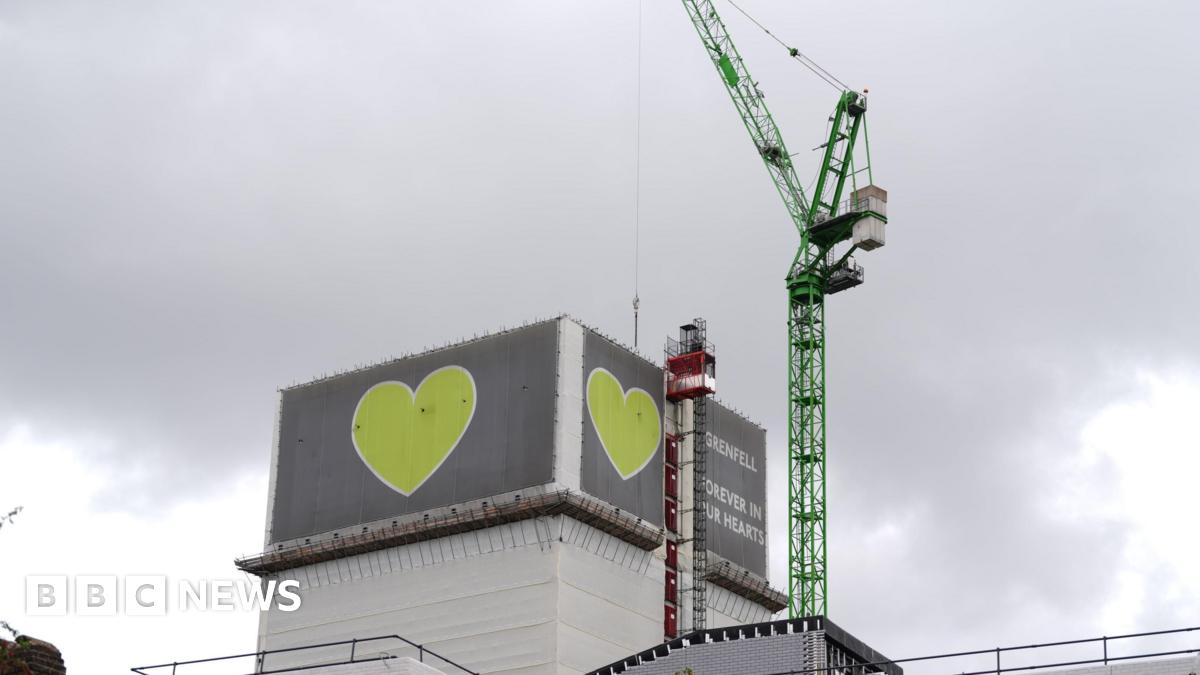 Grenfell Tower partially covered in grey cladding with green heart symbols, as a crane stands beside the building during deconstruction work.
