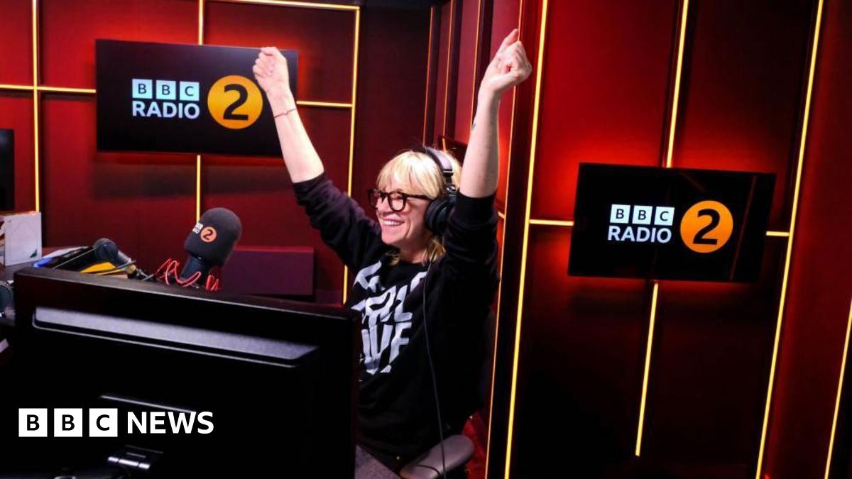 Zoe Ball seen in the BBC Radio 2 studio smiling with her arms raised.