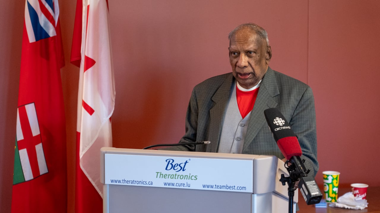 A man standing at a lectern with a CBC microphone in the foreground and two flags in the background