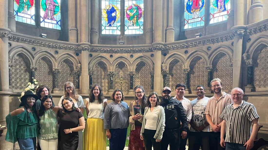 A group of 14 people stands together in a church. Stained-glass windows are visible above them in the background.