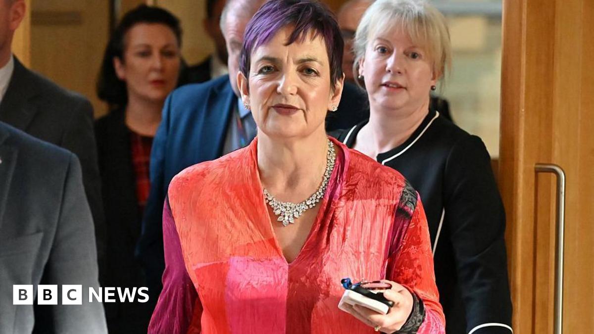Angela Constance, who has short purple-dyed hair and is wearing a red shirt, is walking through the Holyrood building, flanked by Finance Minister Shona Robison and some men in grey suits.