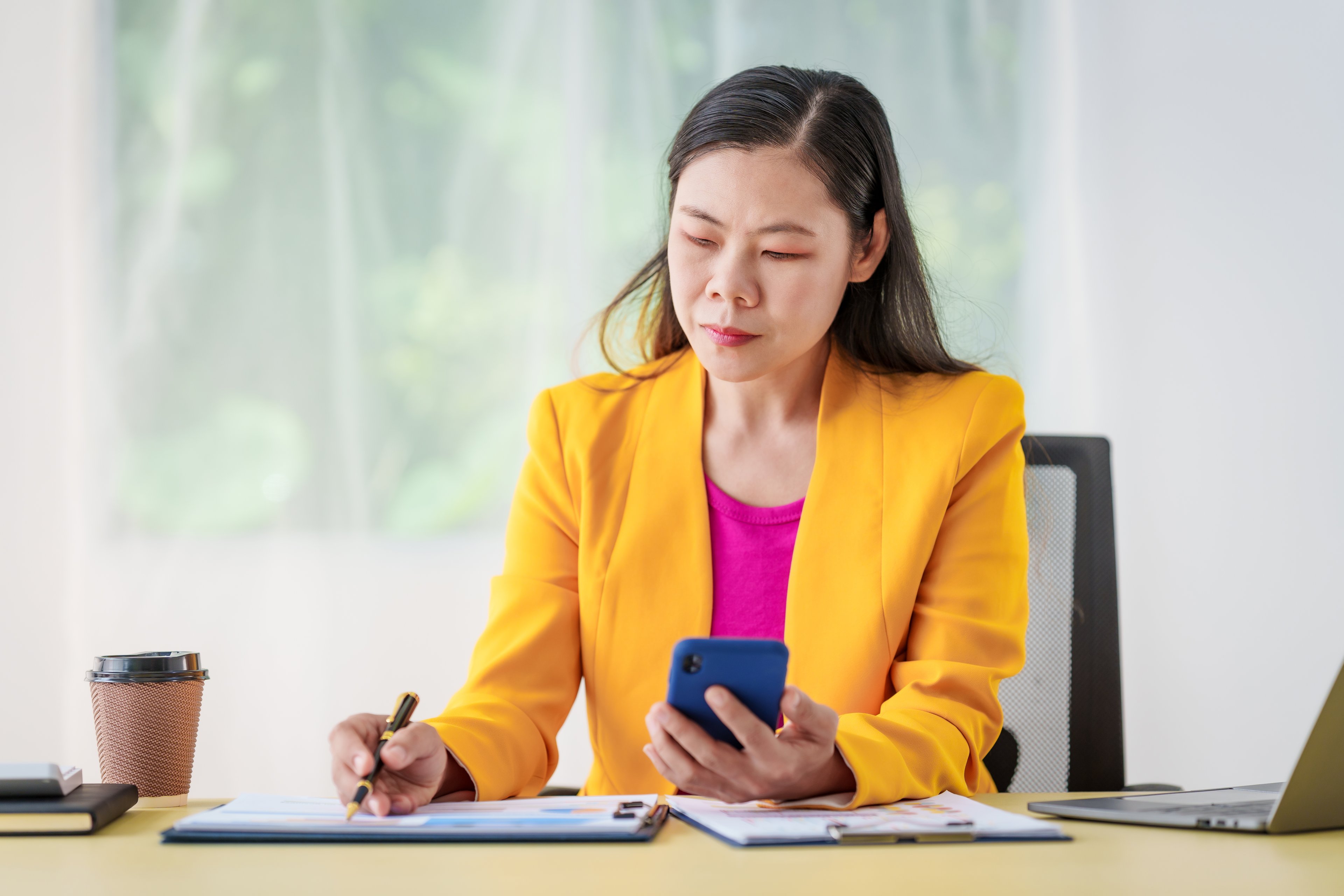 Person at office desk, looking at phone and writing on clipboard.